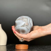 Hand holding a polished agate sphere with clear, gray, and brown quartz formations; carob wood base below.