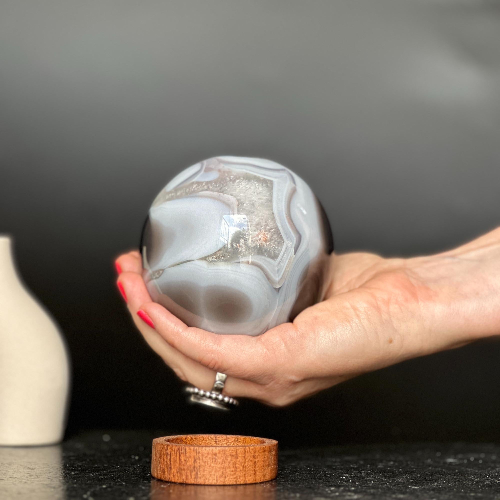 Hand holding a polished agate sphere with clear, gray, and brown quartz formations; carob wood base below.