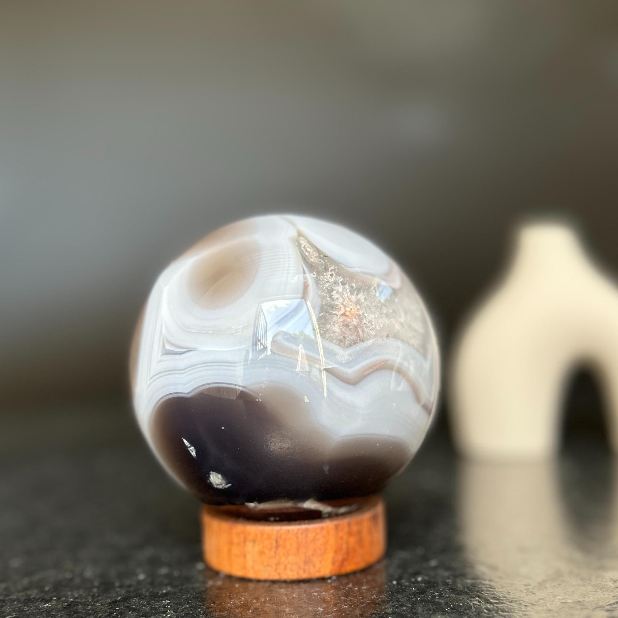 Polished agate sphere with clear, gray, and brown bands, quartz formations, on a round carob wood base.