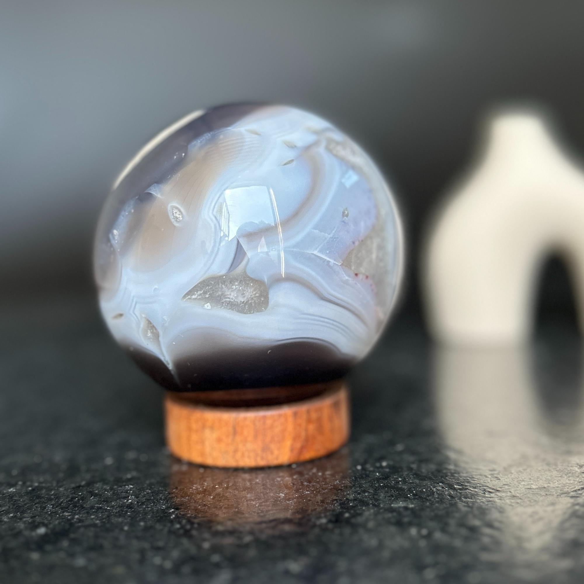 Polished agate sphere with clear, gray, and brown quartz formations on a carob wood base.