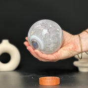 Polished white and beige agate and quartz gemstone sphere, 3.5-inch diameter, held in a hand, with a carob wood base.