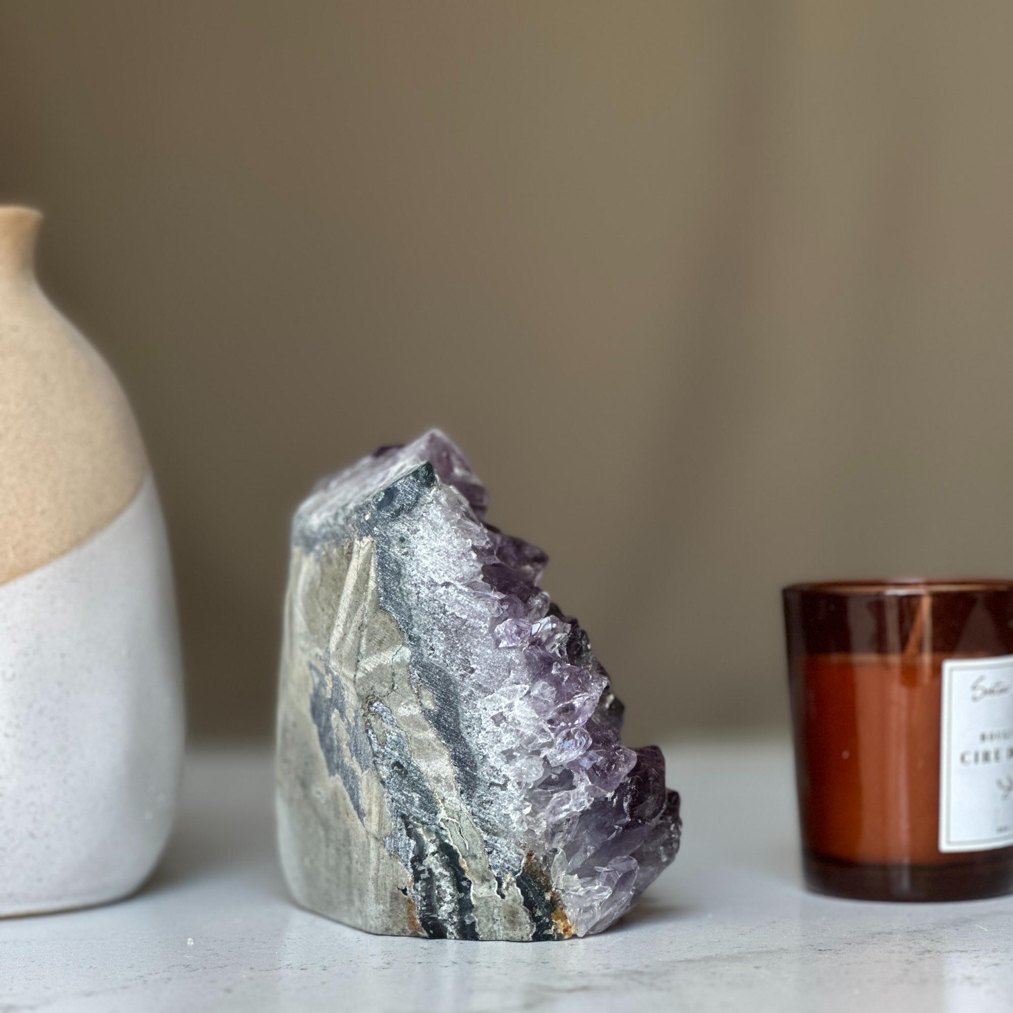 Large, deep purple amethyst geode cluster with a gray and white rock base, next to a two-toned vase and candle.
