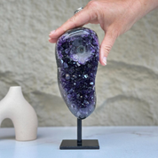 Hand holding a large, self-standing amethyst geode with deep purple, sparkly crystals on a black stand.
