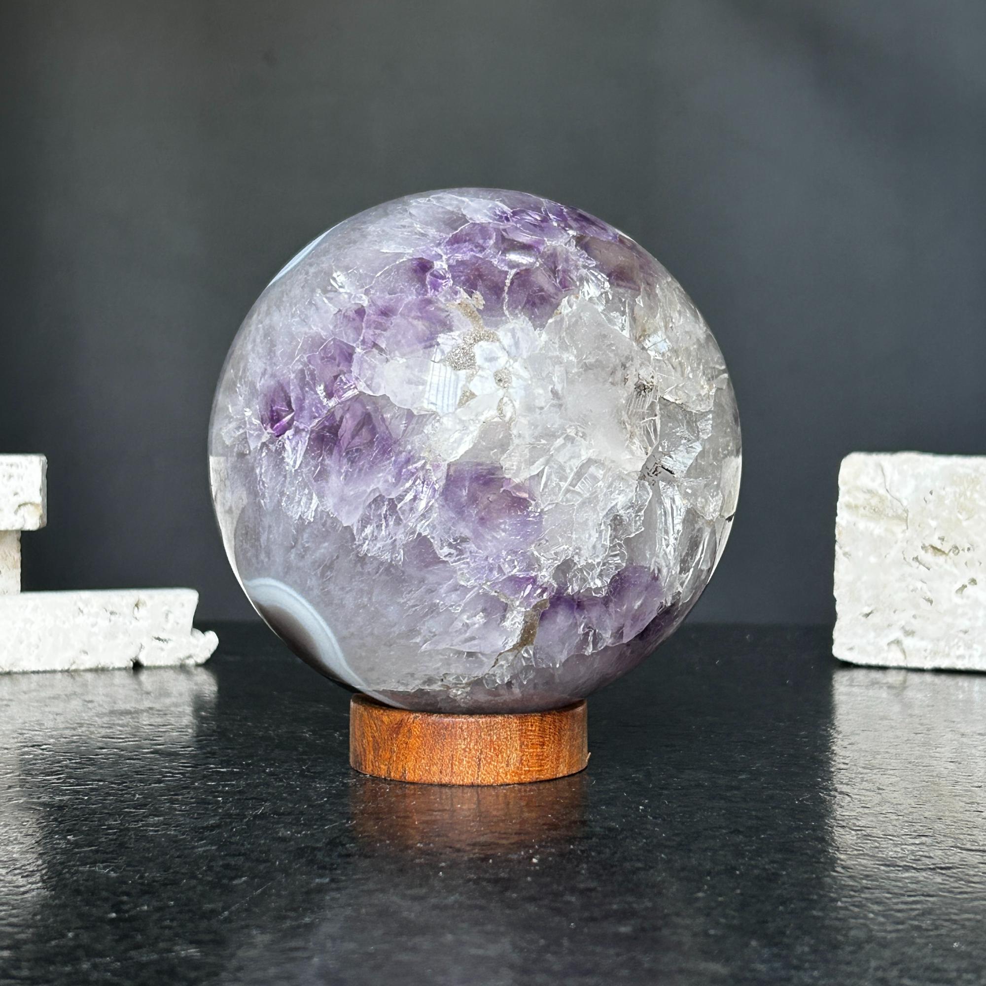 Polished amethyst and agate crystal sphere on a carob wood base, showing purple, gray, and white formations.