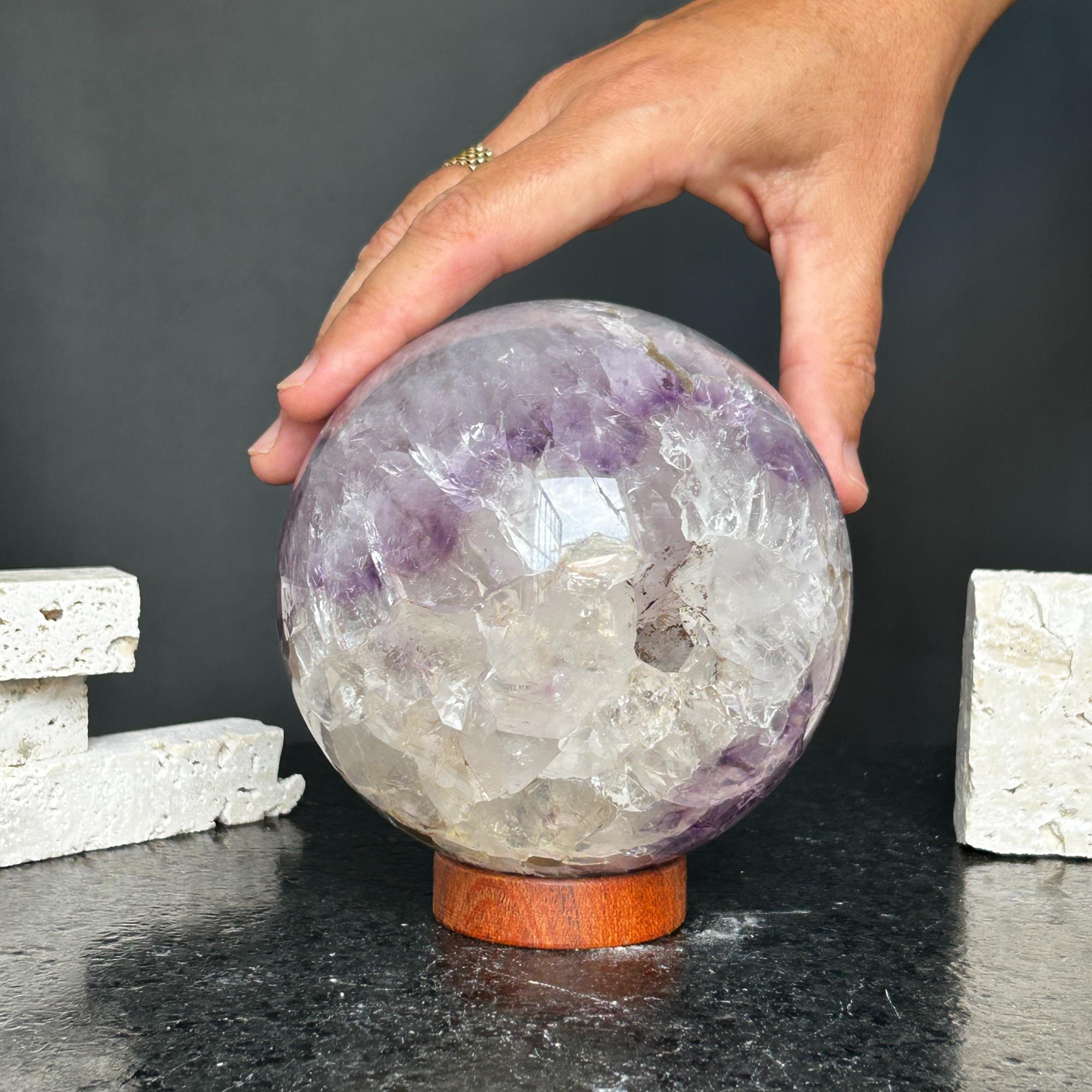 Hand holding a 4-inch amethyst and agate sphere on a carob wood base, showing purple and white crystals.