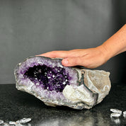 A large amethyst geode with agate formations, displaying vibrant deep purple crystals inside a grey cave-like structure, held by a person.