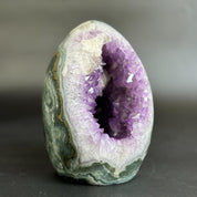 A polished geode specimen with a purple crystal cavity and green-gray banded outer shell, photographed on a dark gray background.