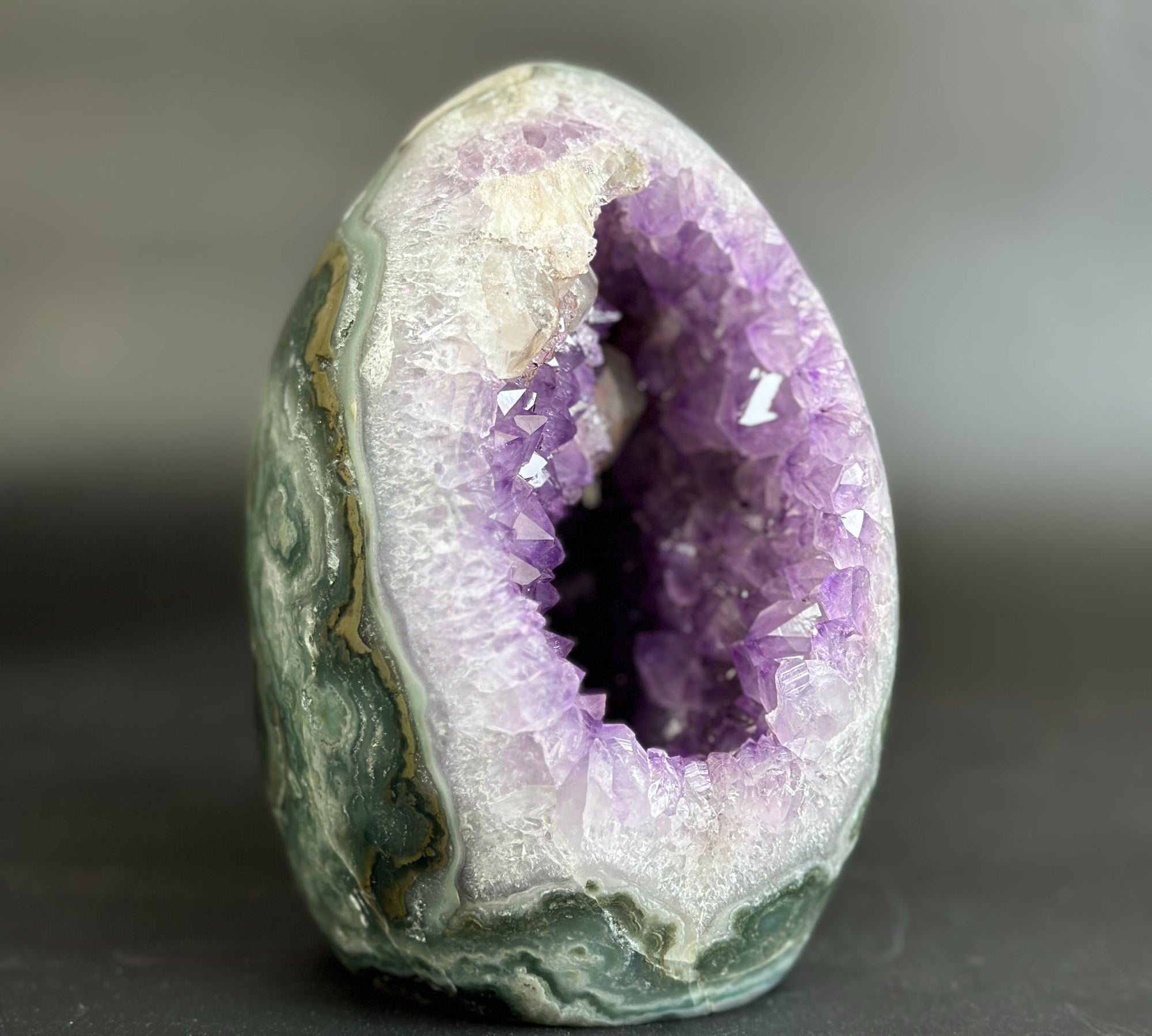 A polished geode specimen with a purple crystal cavity and green-gray banded outer shell, photographed on a dark gray background.