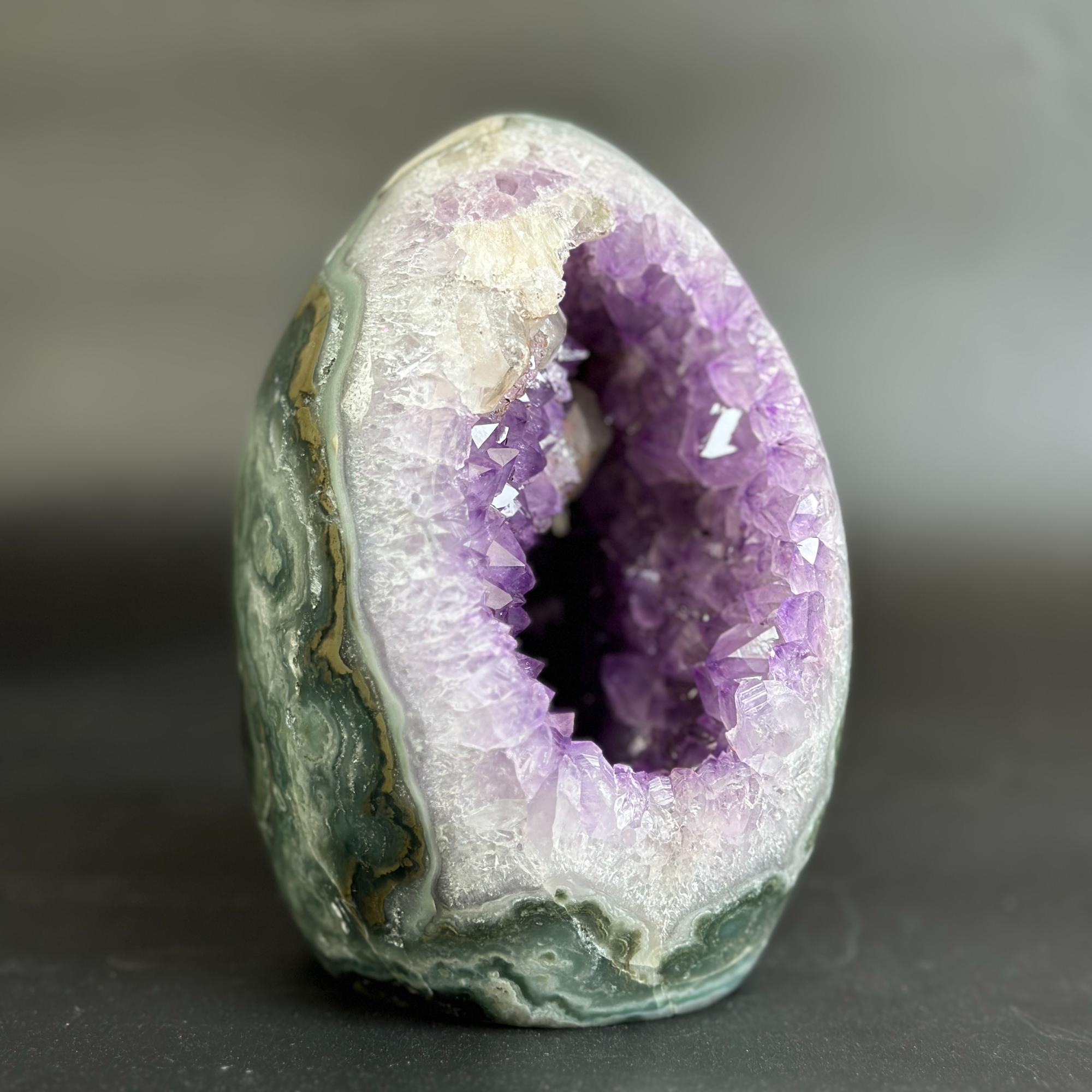 A polished geode specimen with a purple crystal cavity and green-gray banded outer shell, photographed on a dark gray background.