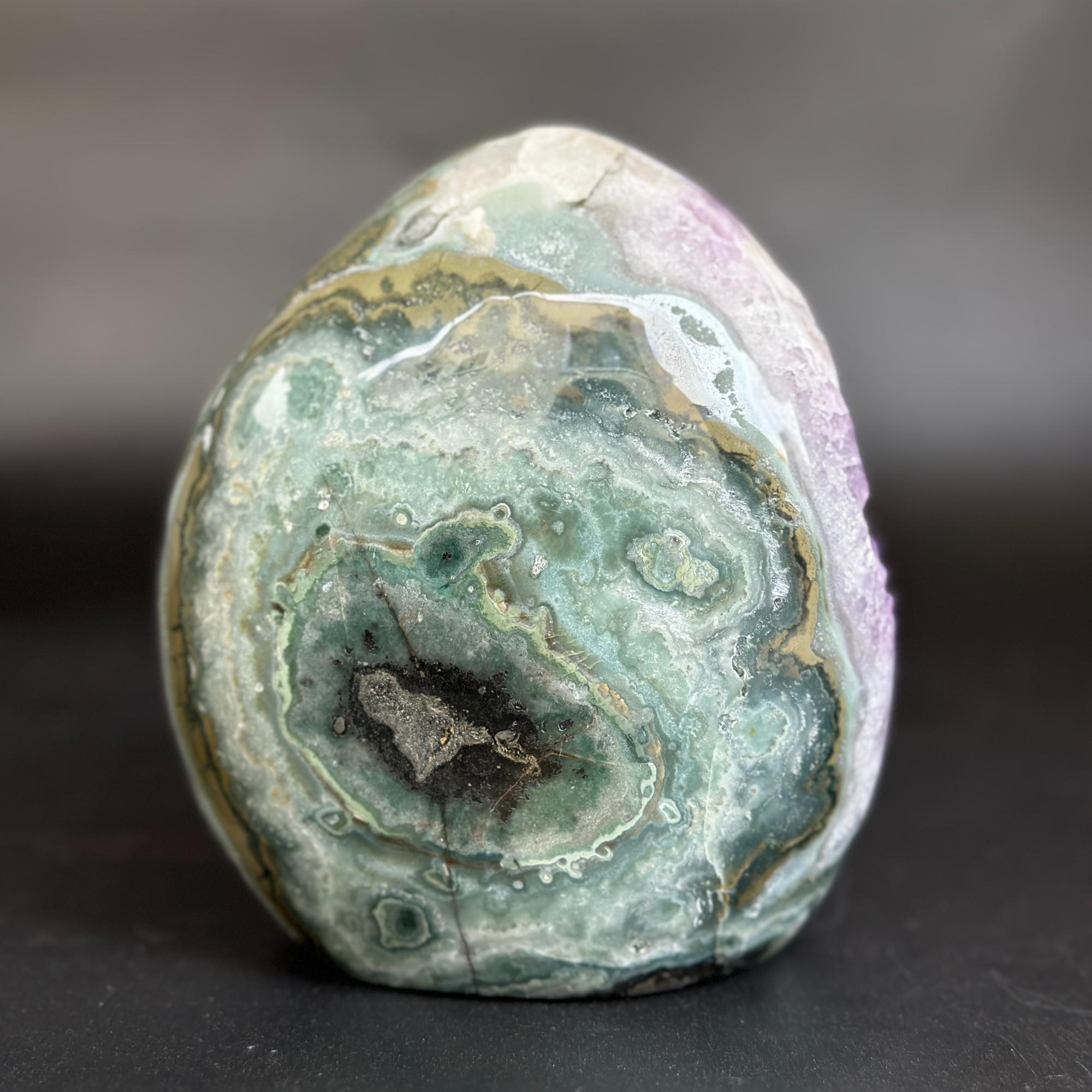 A polished natural stone or agate-like specimen with concentric bands of pale green, white, gray, and lavender-purple, photographed on a dark gray surface against a blurred gray background.
