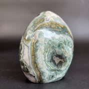 A polished natural stone with concentric bands of green, gray, white, and gold, standing upright on a dark surface against a blurred gray background.