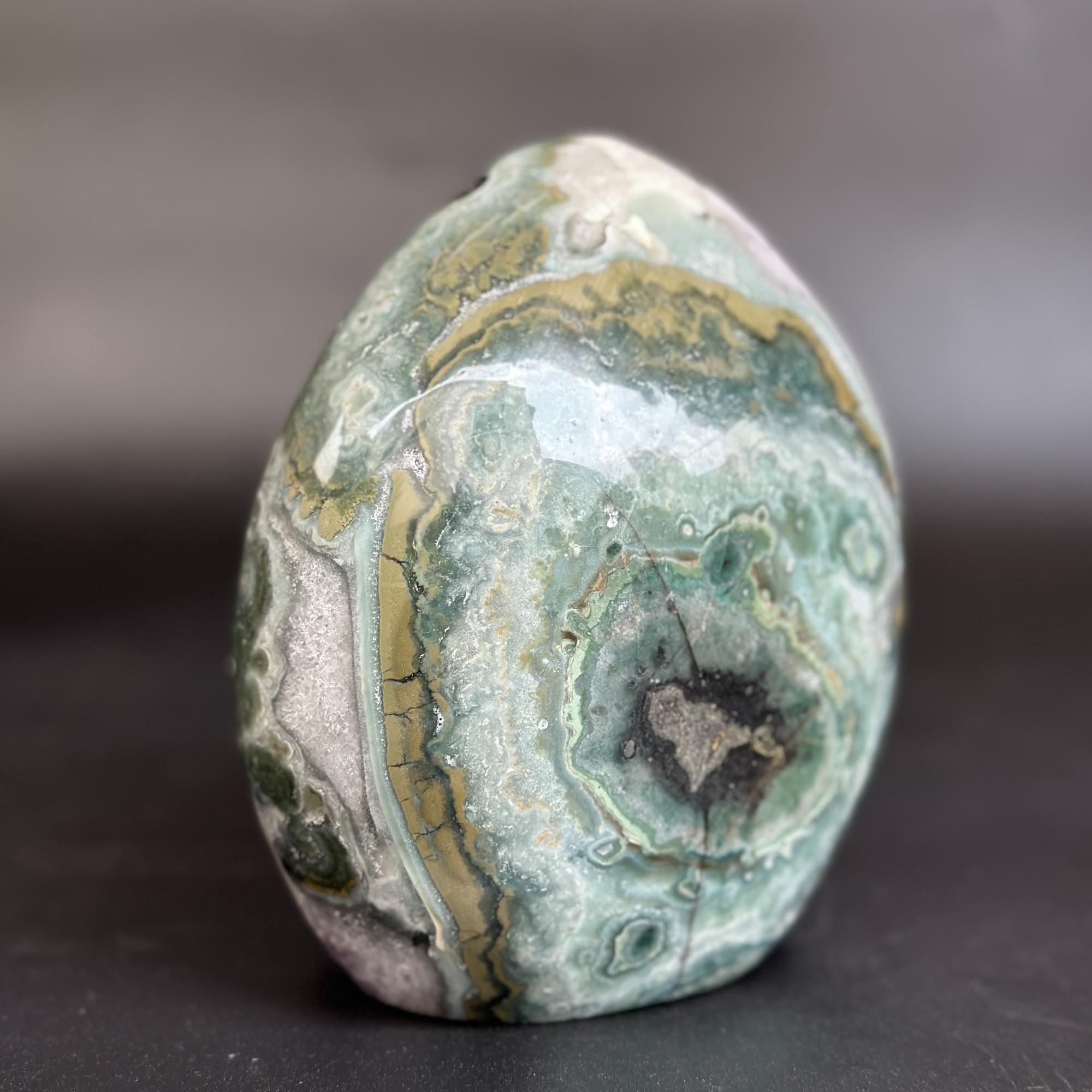 A polished natural stone with concentric bands of green, gray, white, and gold, standing upright on a dark surface against a blurred gray background.