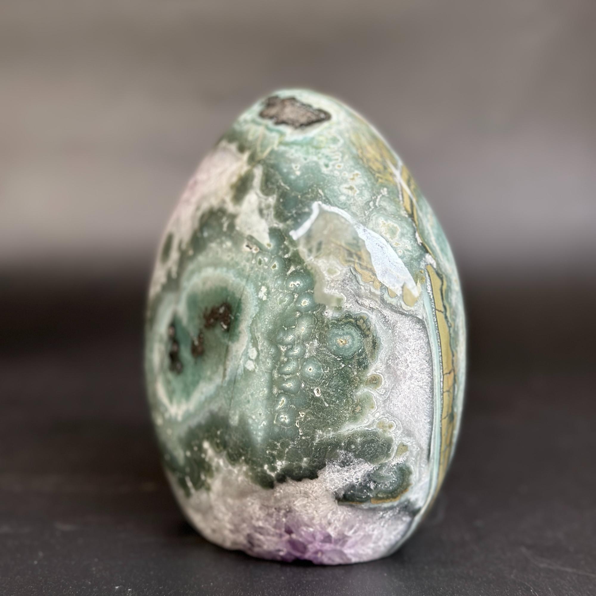 A polished agate crystal stone with swirling patterns of green, white, purple, and gold, standing upright on a dark surface against a blurred gray background.