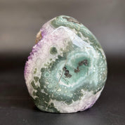 A polished natural stone with swirling bands of green, white, purple, and gray, photographed on a dark surface against a blurred gray background.