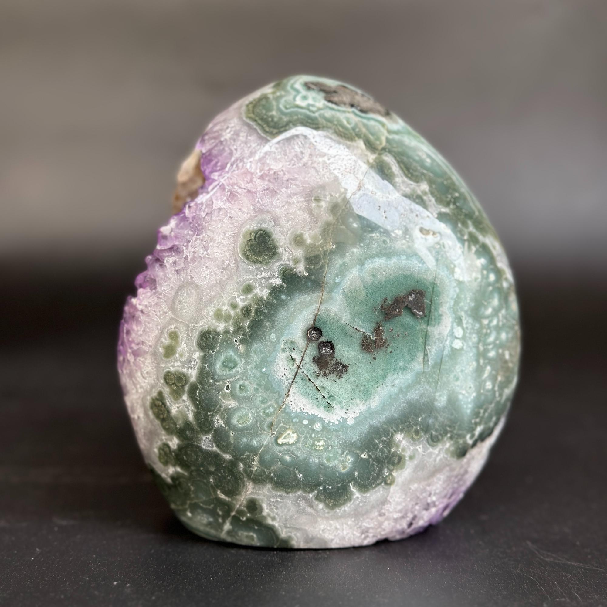 A polished natural stone with swirling bands of green, white, purple, and gray, photographed on a dark surface against a blurred gray background.