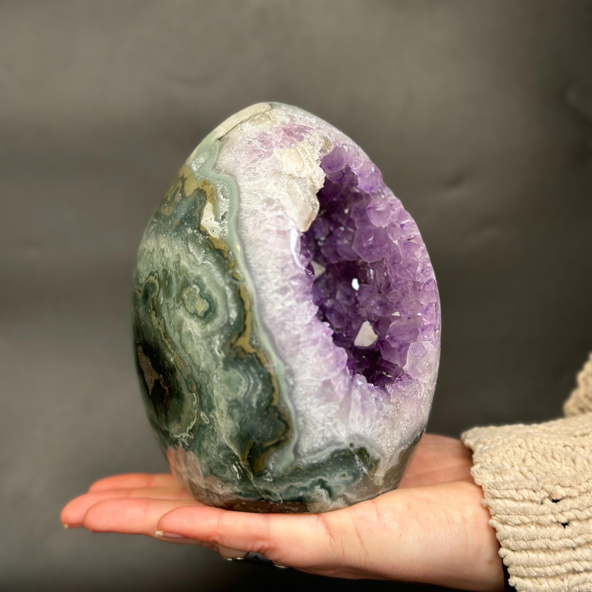 A polished amethyst geode with green and white agate patterns, held in a person's hand against a dark gray background.