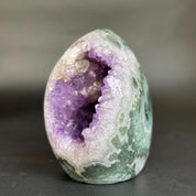 A polished geode specimen with a purple crystal cavity and green-and-white marbled outer shell, photographed on a dark neutral surface.