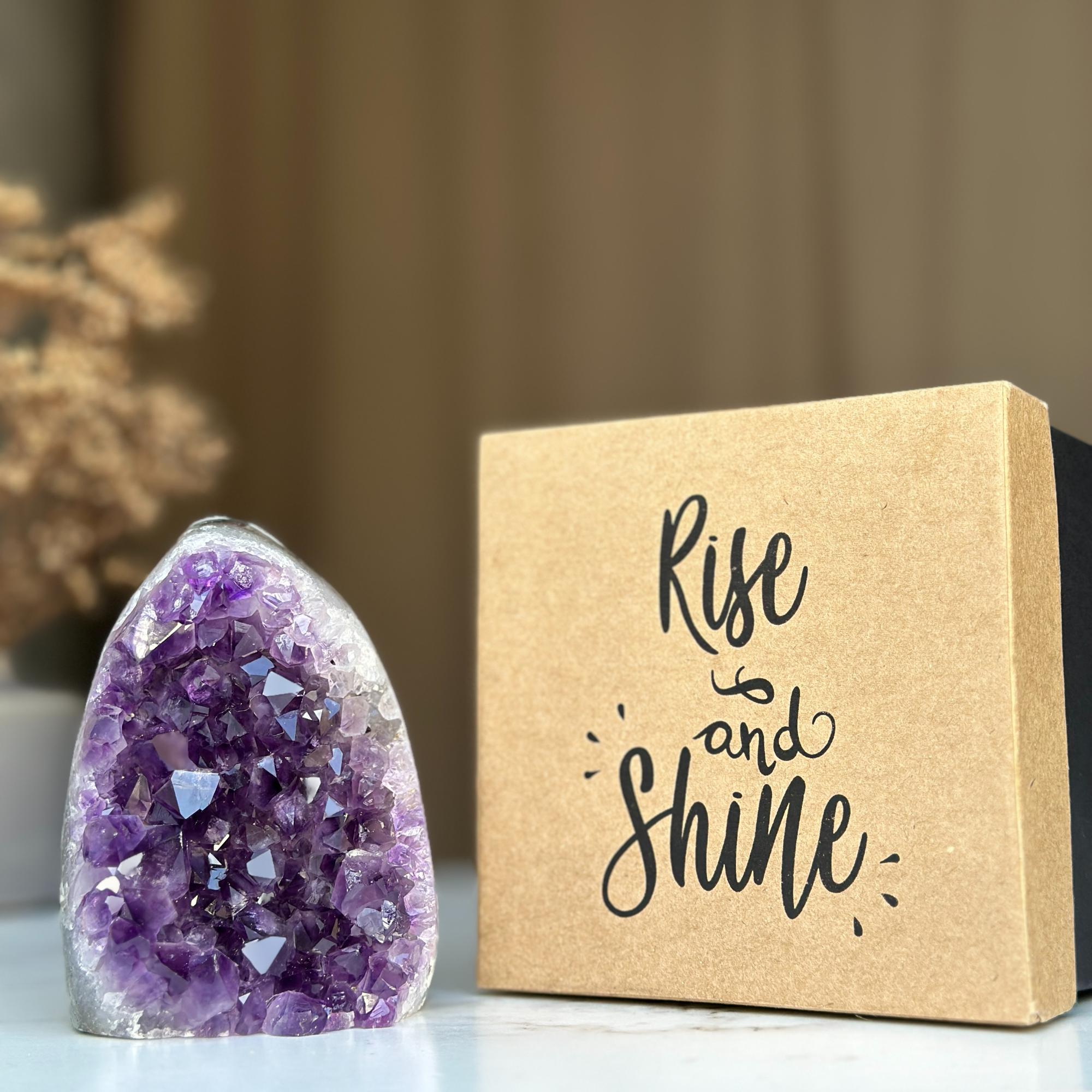 Deep purple amethyst geode next to a premium brown kraft gift box with 'Rise and Shine' script.