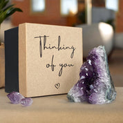 Vibrant purple amethyst geode and small crystals next to a kraft brown and black "Thinking of you" gift box.