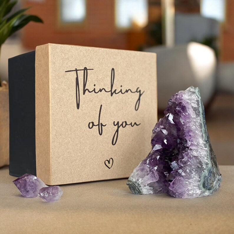 Vibrant purple amethyst geode and small crystals next to a kraft brown and black "Thinking of you" gift box.