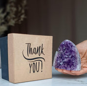 Two-tone kraft and dark rigid gift box with "Thank YOU!" script, beside a hand holding a sparkling purple amethyst geode.