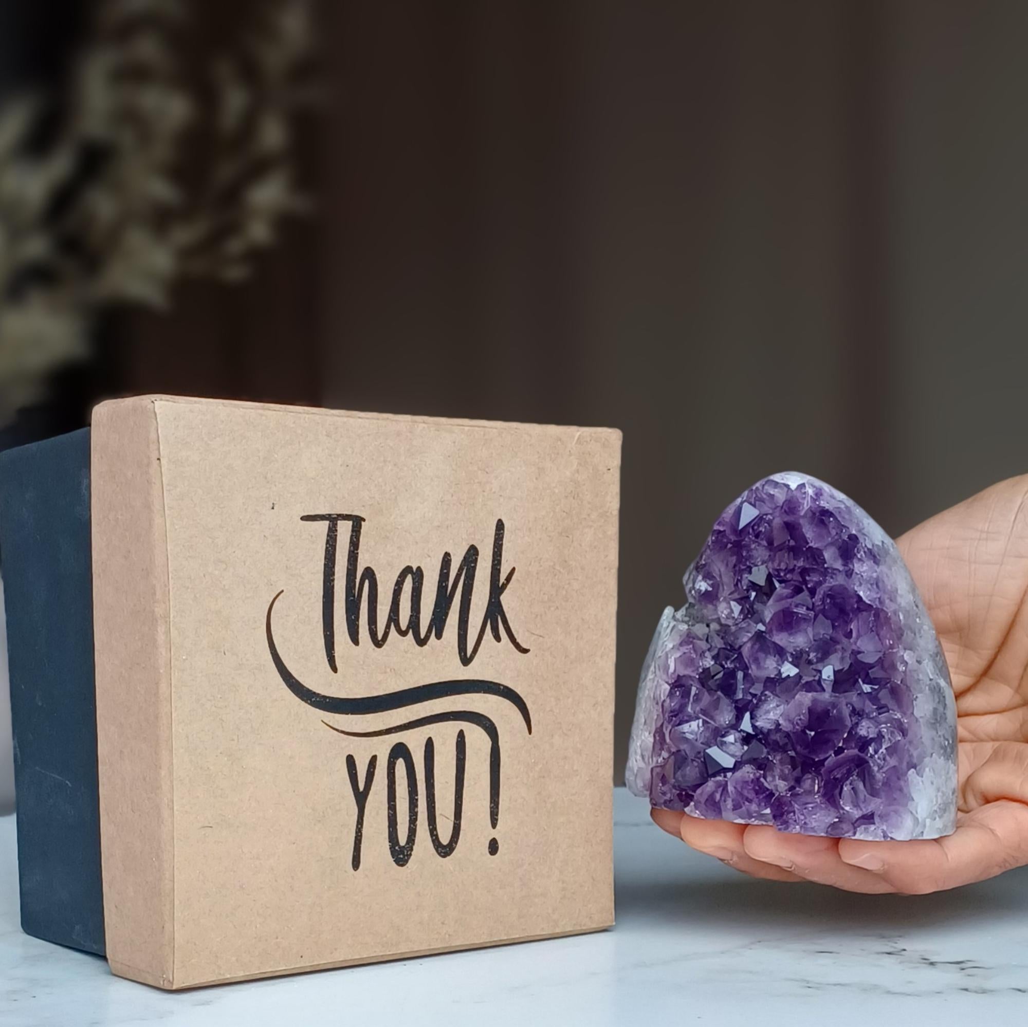 Two-tone kraft and dark rigid gift box with "Thank YOU!" script, beside a hand holding a sparkling purple amethyst geode.