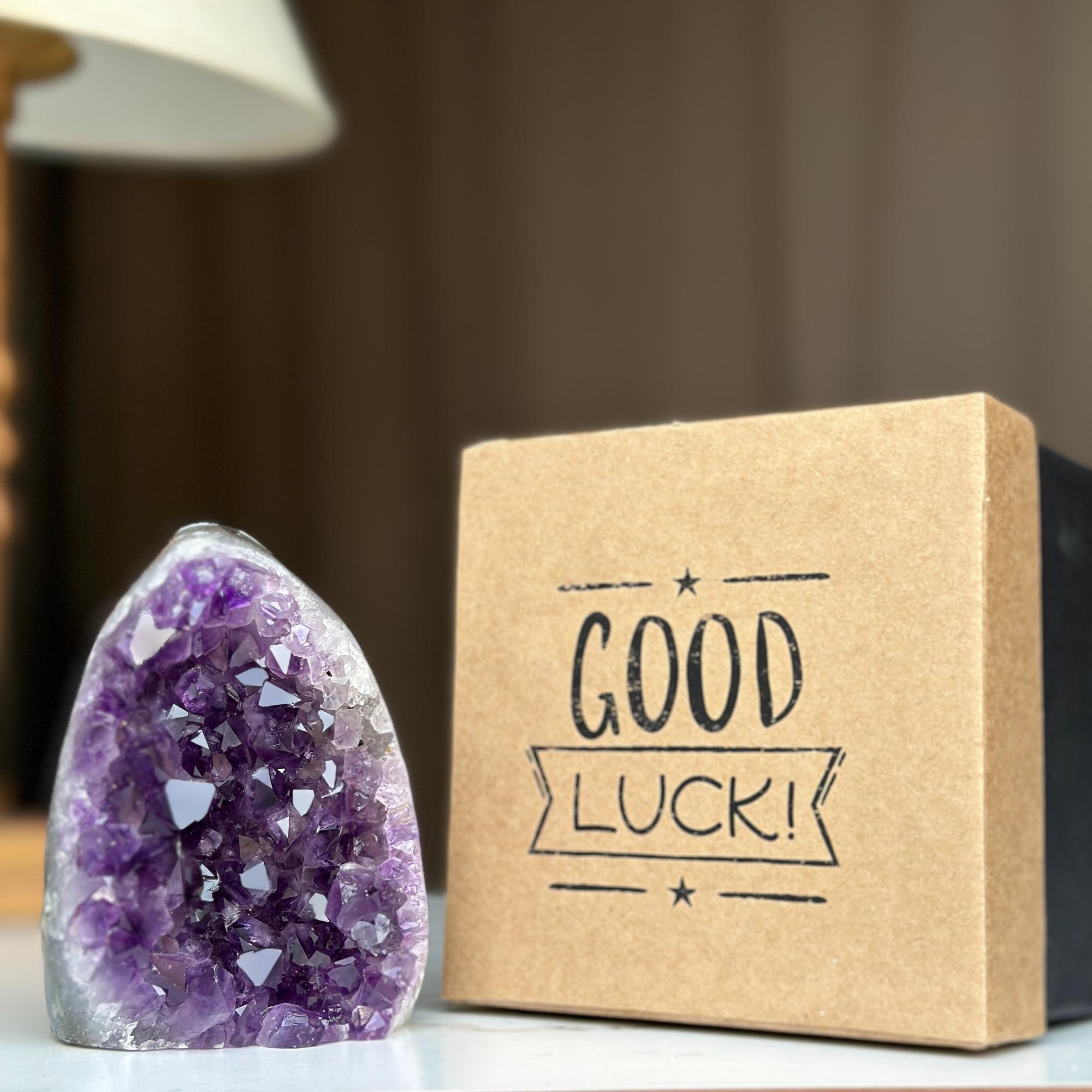 Vibrant purple amethyst geode next to a rigid kraft brown gift box with "GOOD LUCK!" text. Premium anniversary gift.