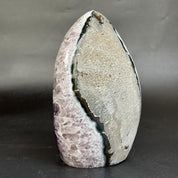 A polished natural stone sculpture with a teardrop shape, featuring a prominent vertical fissure. The left side of the stone displays a lavender-purple crystalline texture, while the right side is a textured light gray and beige matrix.
