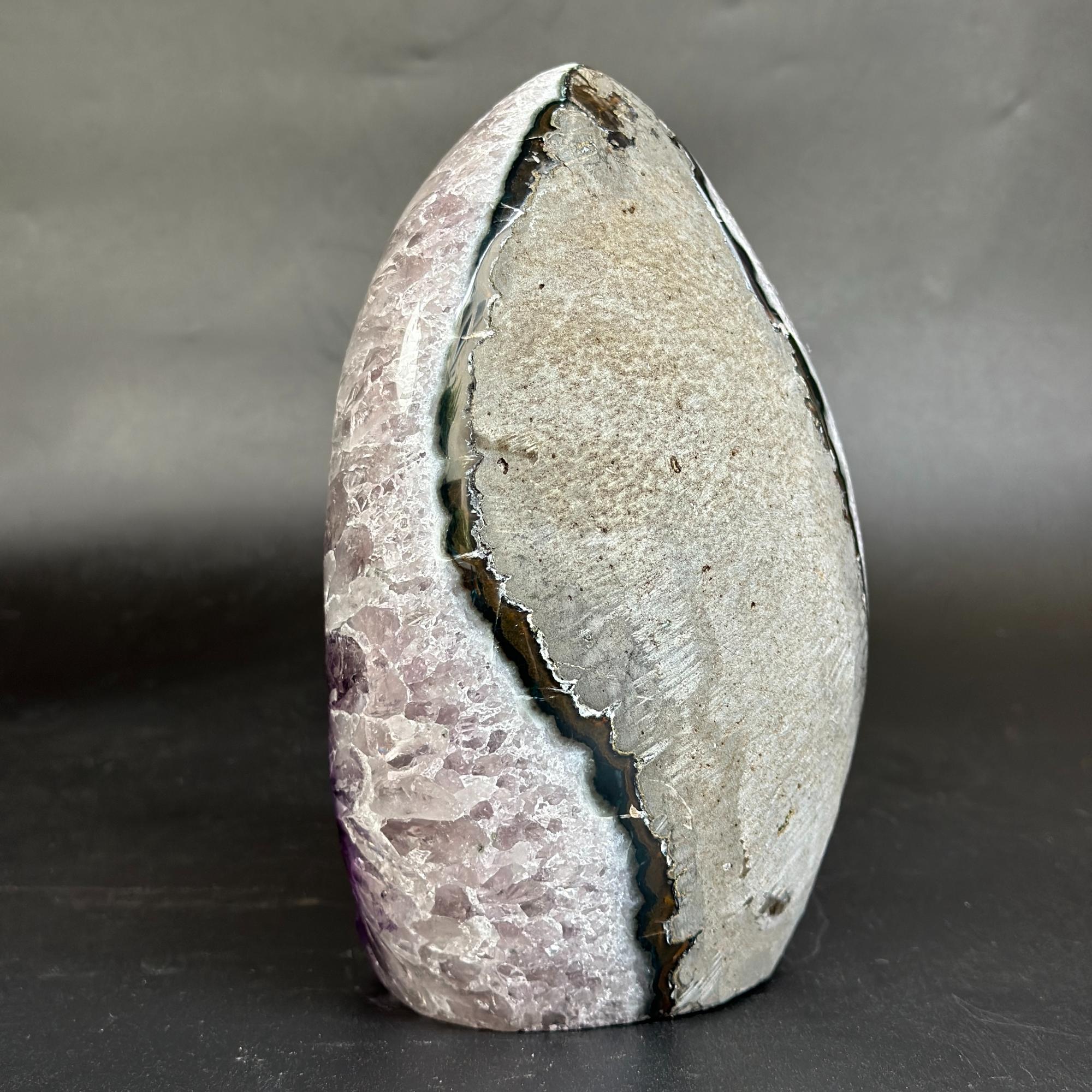 A polished natural stone sculpture with a teardrop shape, featuring a prominent vertical fissure. The left side of the stone displays a lavender-purple crystalline texture, while the right side is a textured light gray and beige matrix.