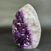 A natural amethyst crystal geode specimen with a deep purple crystal cavity and a clear, translucent white outer matrix, standing upright on a dark surface against a blurred gray background.