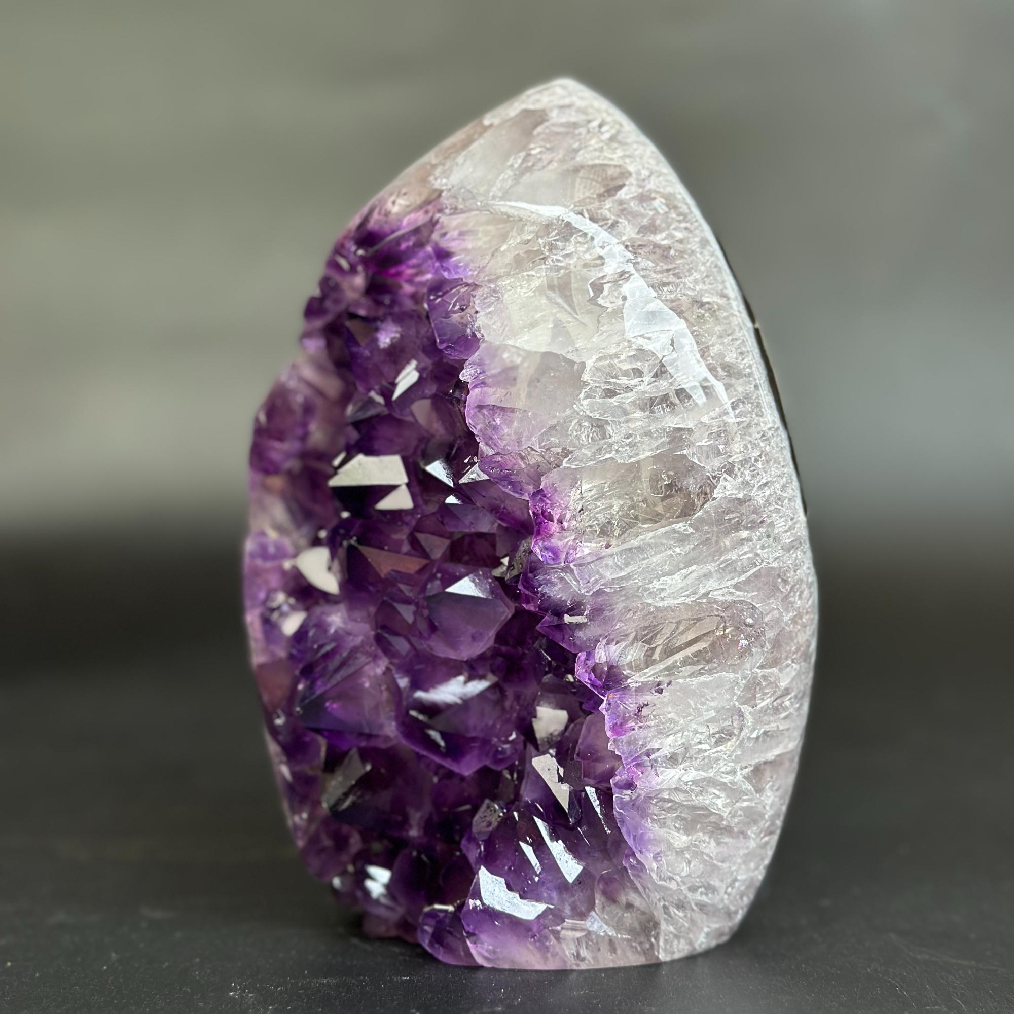 A natural amethyst crystal geode specimen with a deep purple crystal cavity and a clear, translucent white outer matrix, standing upright on a dark surface against a blurred gray background.