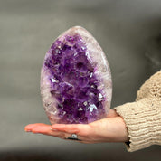 A person's hand holding a large, polished amethyst geode crystal with deep purple and white crystalline structures, set against a soft, blurred gray background.