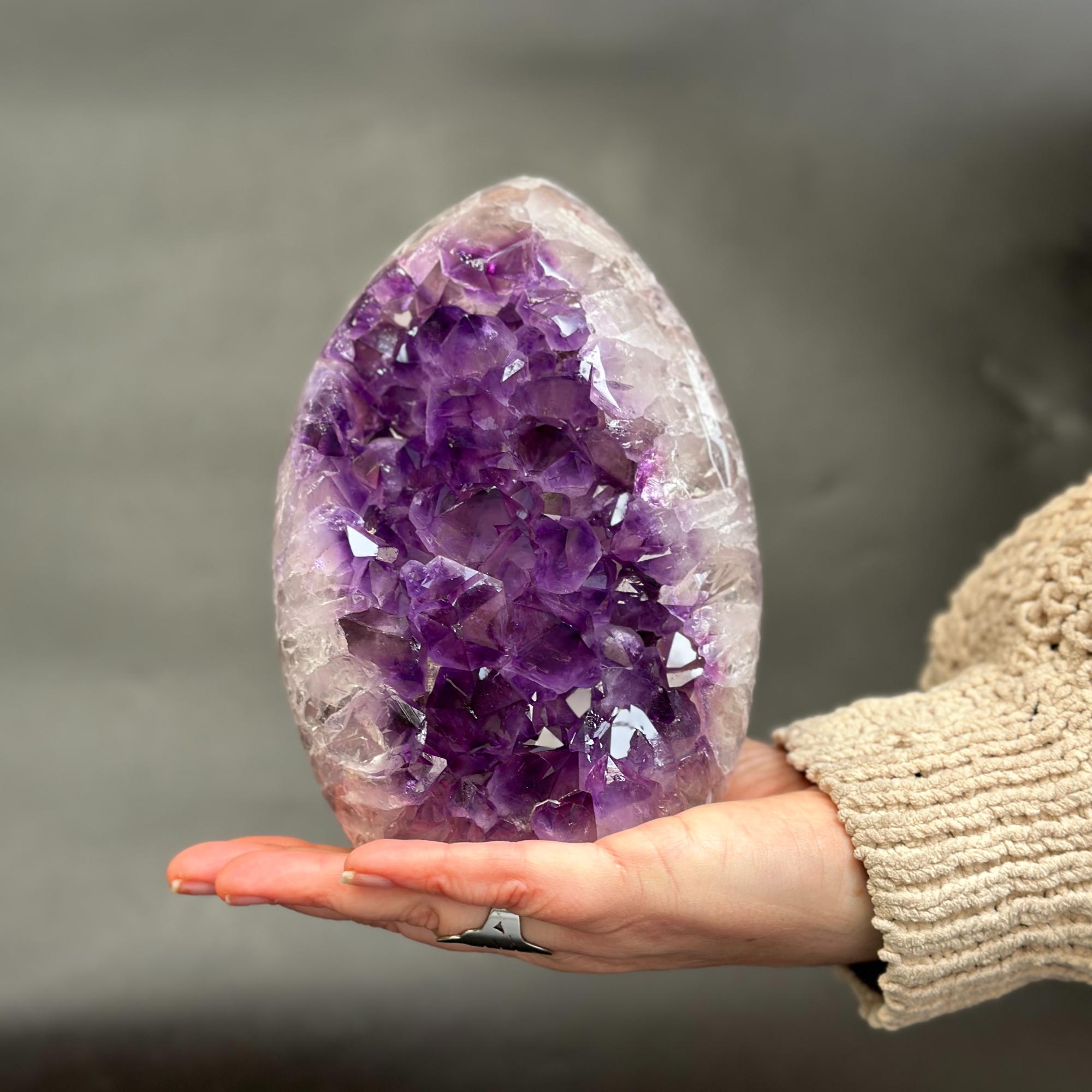A person's hand holding a large, polished amethyst geode crystal with deep purple and white crystalline structures, set against a soft, blurred gray background.