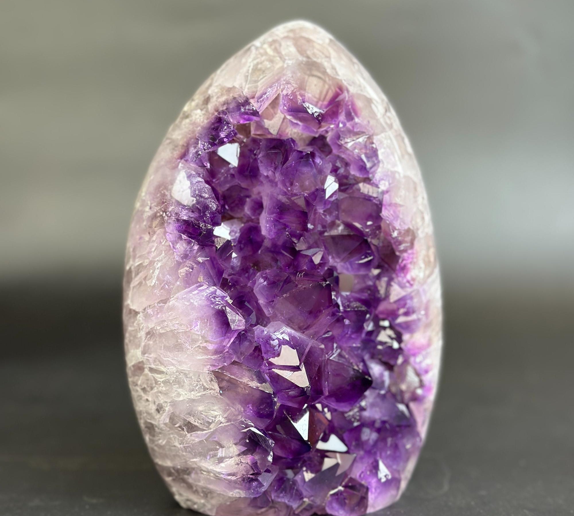 A polished amethyst crystal geode specimen with a pointed oval shape, featuring a dense cluster of deep purple crystals on one side and a translucent pale lavender to off-white outer matrix on the other, standing upright on a dark surface against a soft gray background.