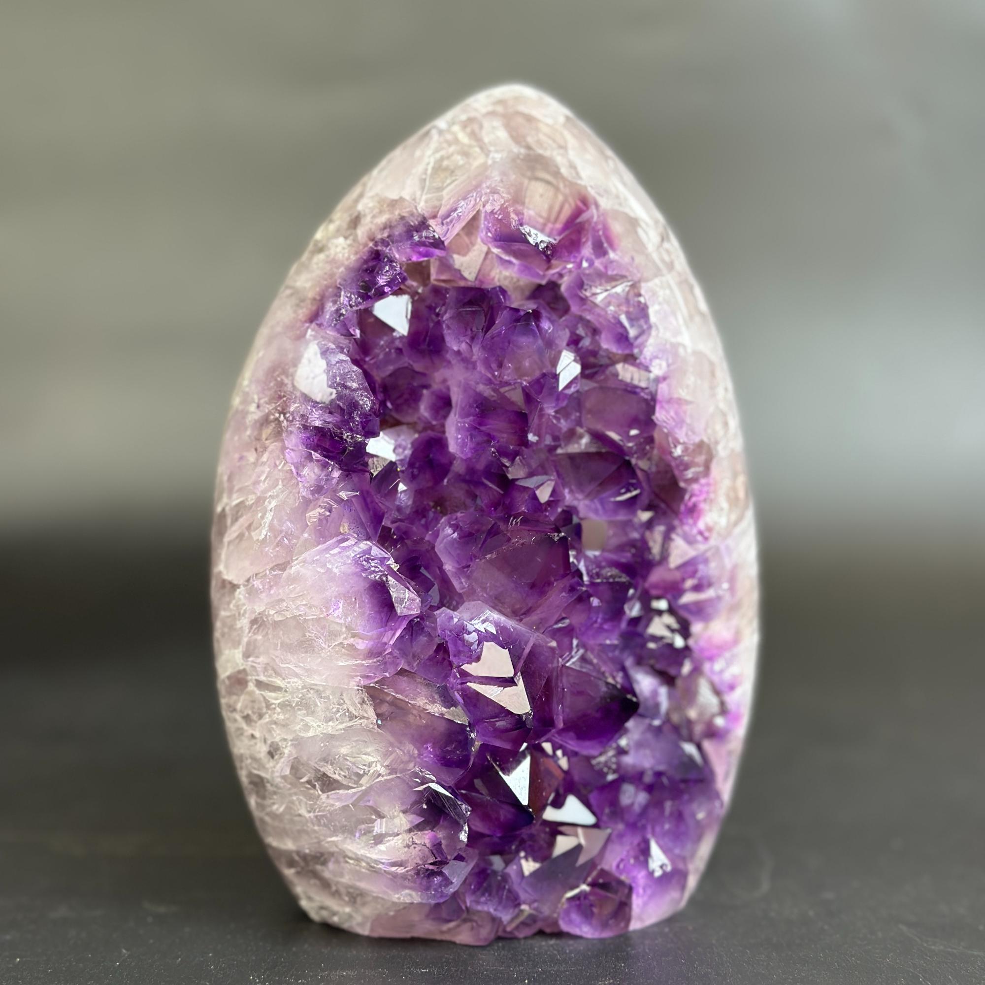 A polished amethyst crystal geode specimen with a pointed oval shape, featuring a dense cluster of deep purple crystals on one side and a translucent pale lavender to off-white outer matrix on the other, standing upright on a dark surface against a soft gray background.