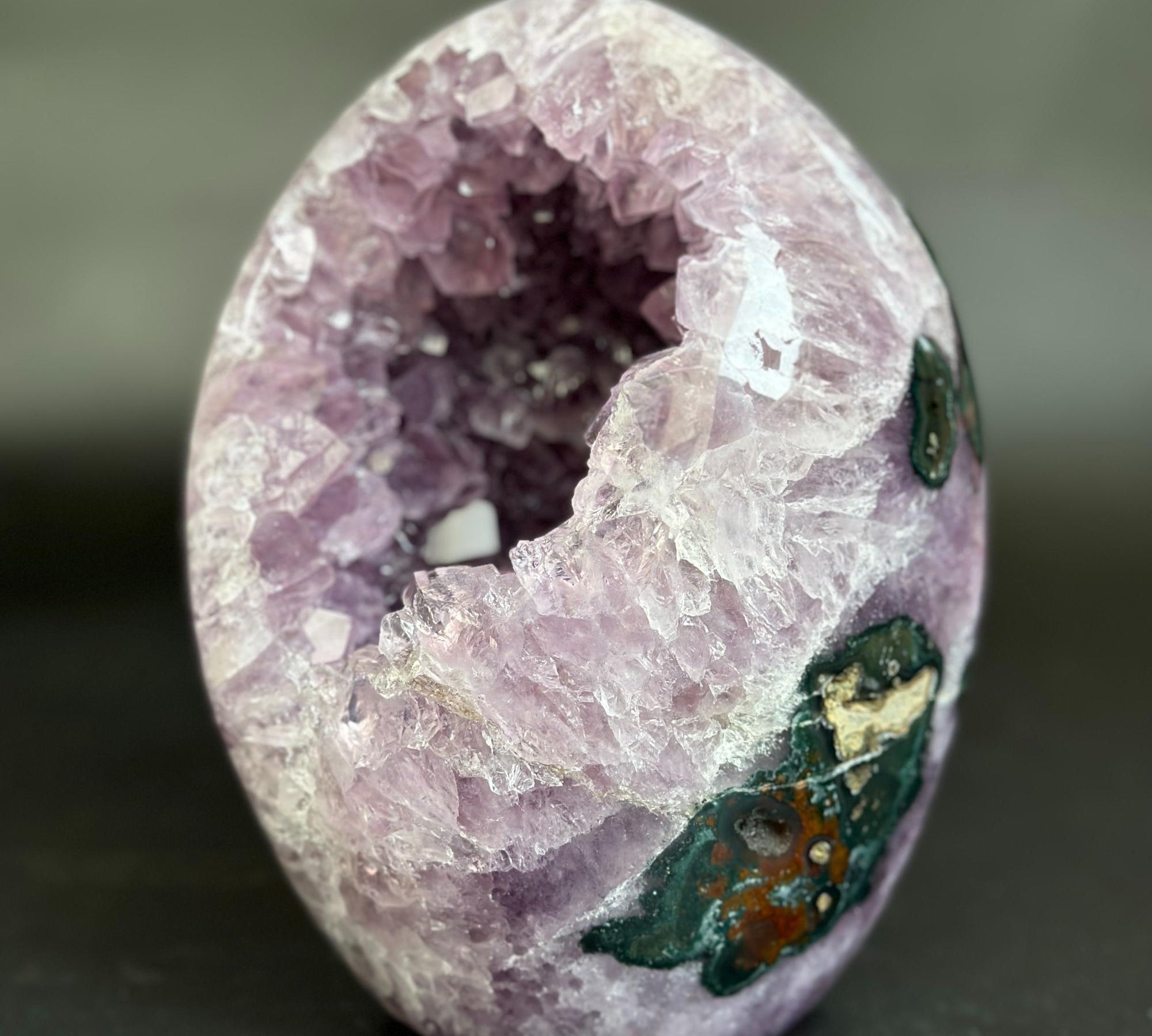 A polished geode specimen with a hollow interior, featuring lavender and purple crystal clusters on one side and dark green, brown, and white mineral inclusions on the outer surface, set against a dark gray background.