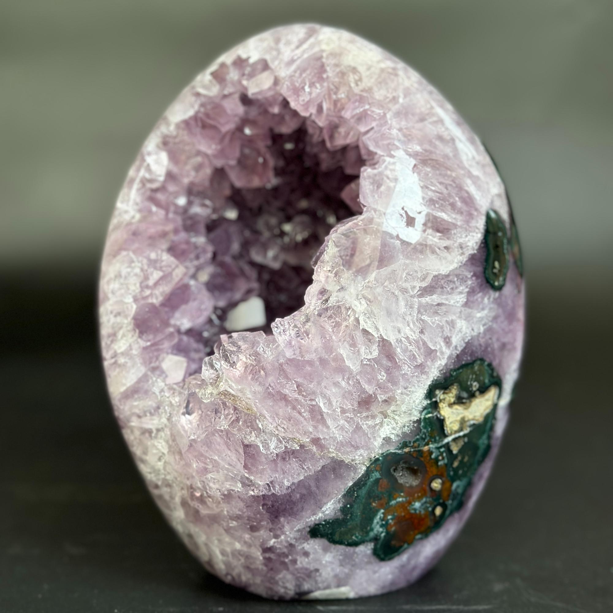A polished geode specimen with a hollow interior, featuring lavender and purple crystal clusters on one side and dark green, brown, and white mineral inclusions on the outer surface, set against a dark gray background.