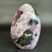 A polished purple amethyst crystal geode with dark green and brown mineral inclusions, photographed on a dark gray background.