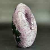 A polished natural crystal geode specimen with a pinkish-purple outer matrix, a hollow white crystalline interior, and dark green mineral inclusions on one side, photographed against a soft gray background.