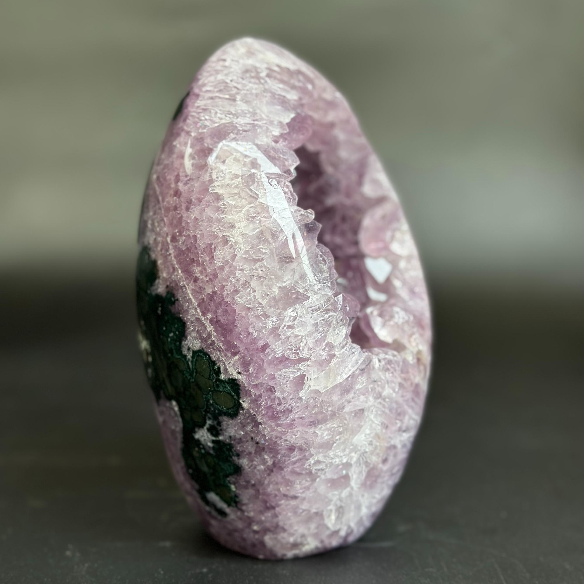 A polished natural crystal geode specimen with a pinkish-purple outer matrix, a hollow white crystalline interior, and dark green mineral inclusions on one side, photographed against a soft gray background.