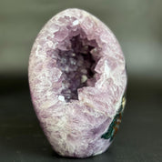 A natural amethyst geode sculpture with a hollow interior, showcasing light purple crystals and a textured, crystalline outer surface, photographed on a dark gray background.