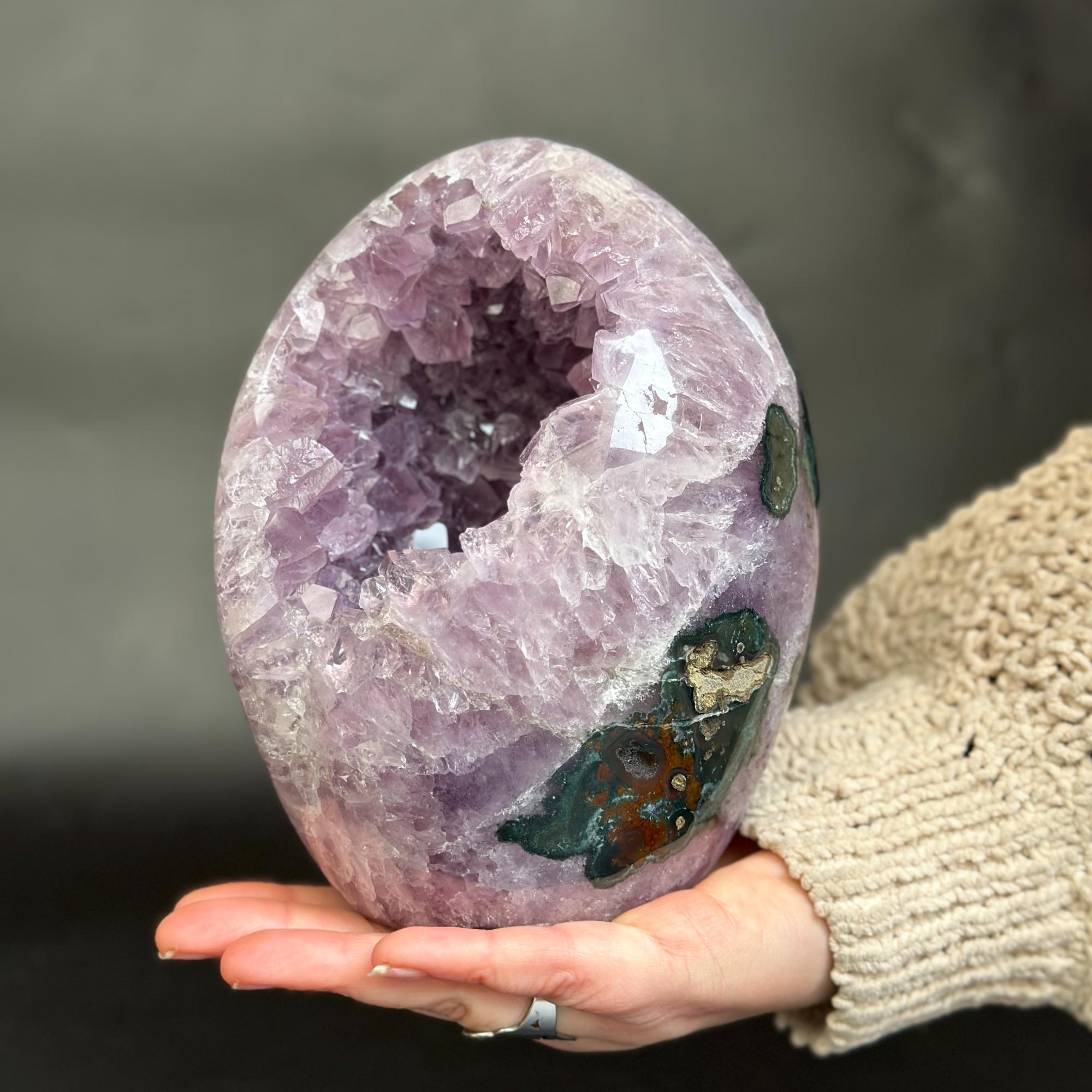 A hand holds a polished purple amethyst geode crystal sphere with a hollow center and natural inclusions, against a dark gray background.