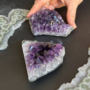 Two deep purple Uruguayan amethyst clusters, one held by a hand, on a dark surface with other crystal geodes.