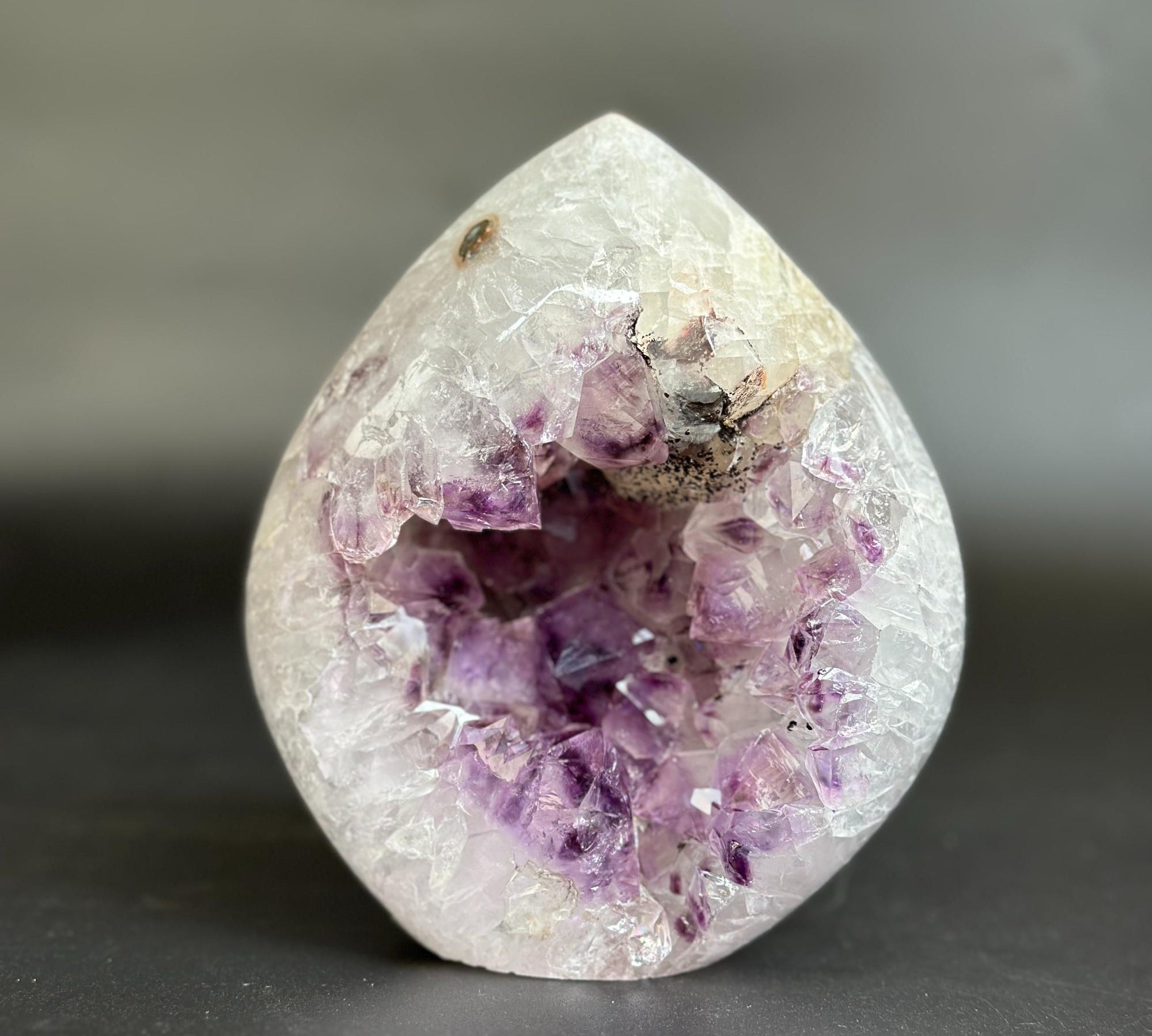 A teardrop-shaped crystal geode sculpture made of clear quartz with a prominent purple amethyst cavity inside, set against a dark gray background.