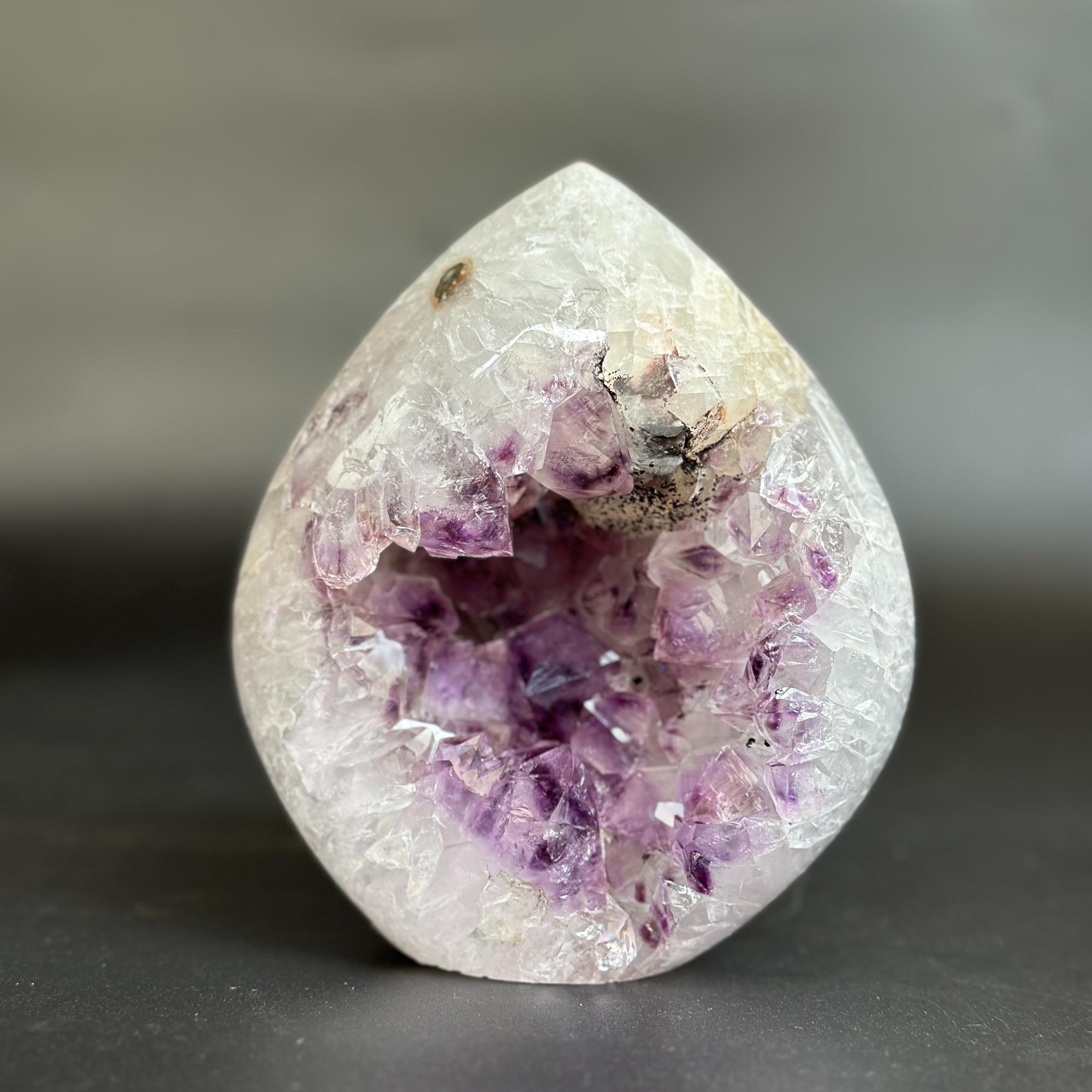 A teardrop-shaped crystal geode sculpture made of clear quartz with a prominent purple amethyst cavity inside, set against a dark gray background.