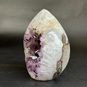 A polished teardrop-shaped crystal geode specimen featuring a white and pale beige outer crystalline structure, a deep purple crystal cavity, and brown matrix details, displayed on a dark surface against a dark gray background.