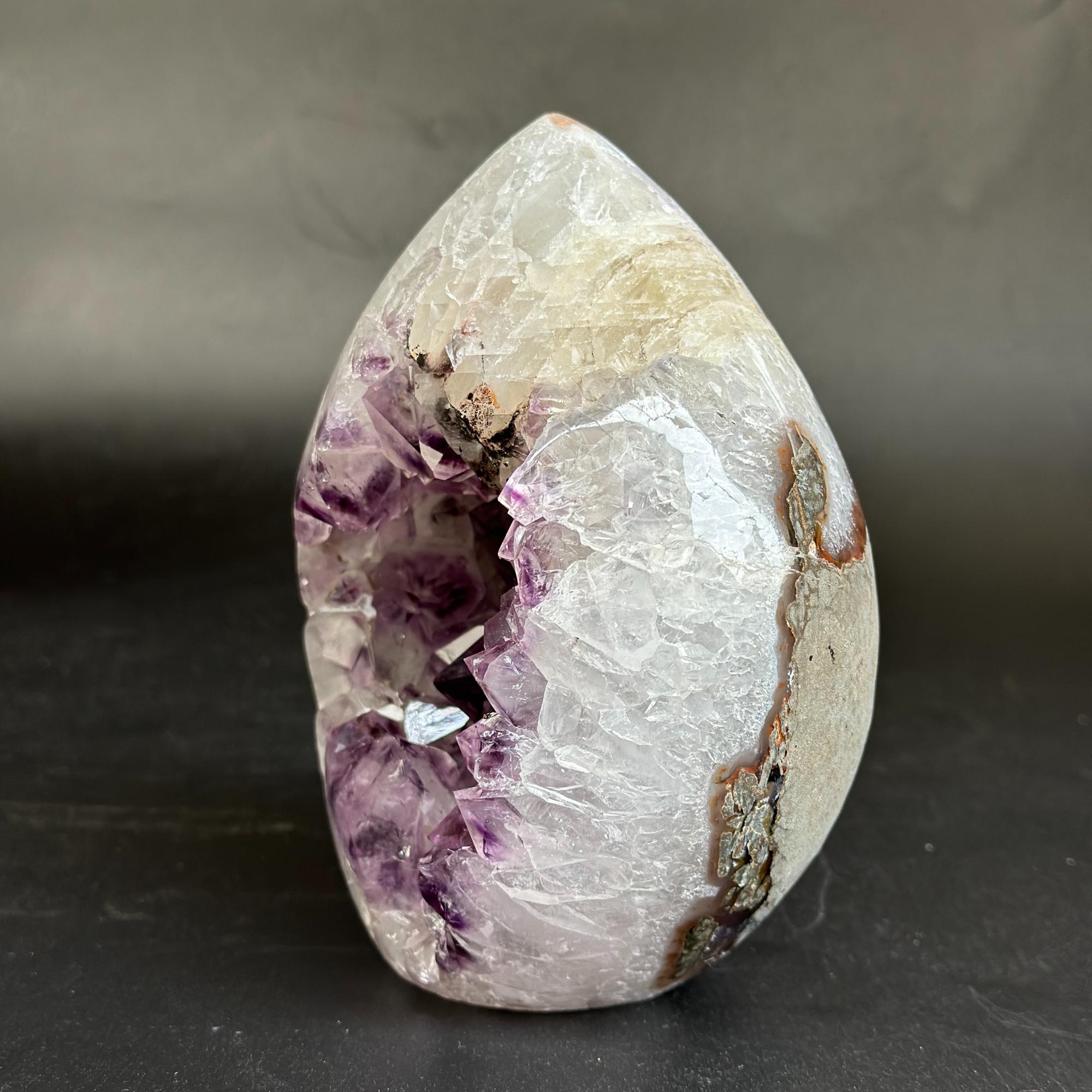 A polished teardrop-shaped crystal geode specimen featuring a white and pale beige outer crystalline structure, a deep purple crystal cavity, and brown matrix details, displayed on a dark surface against a dark gray background.