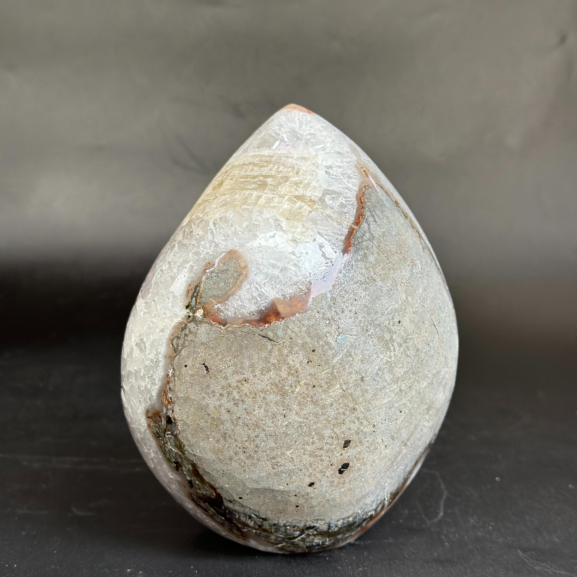 A polished teardrop-shaped natural stone sculpture with a textured, speckled surface in shades of gray, beige, and brown, standing upright on a dark surface against a dark gray background.