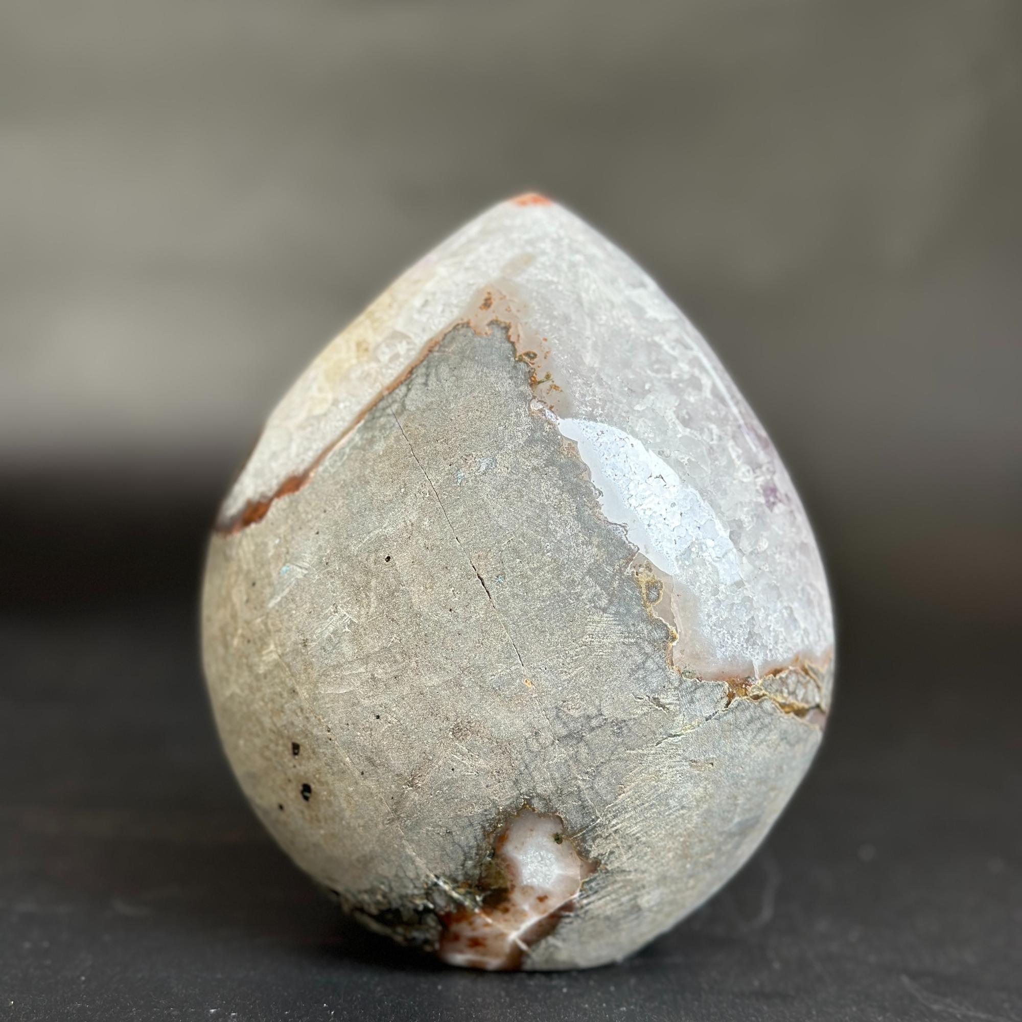 A polished teardrop-shaped natural stone or crystal with a mottled gray, beige, and white surface and subtle brown veining, photographed on a dark surface against a blurred gray background.