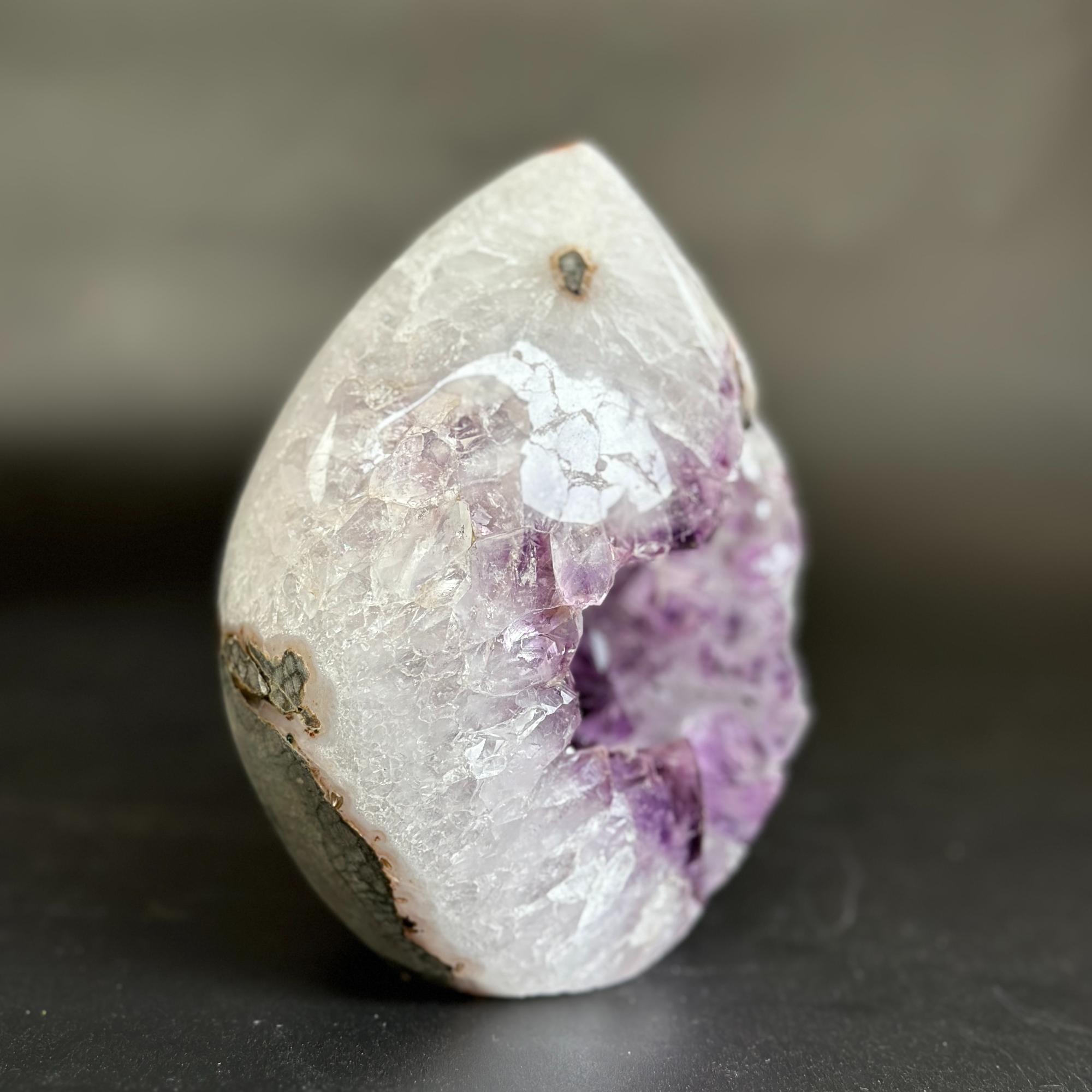 A polished natural crystal geode specimen with a white and lavender-purple interior and a dark gray outer surface, photographed on a dark surface against a blurred gray background.